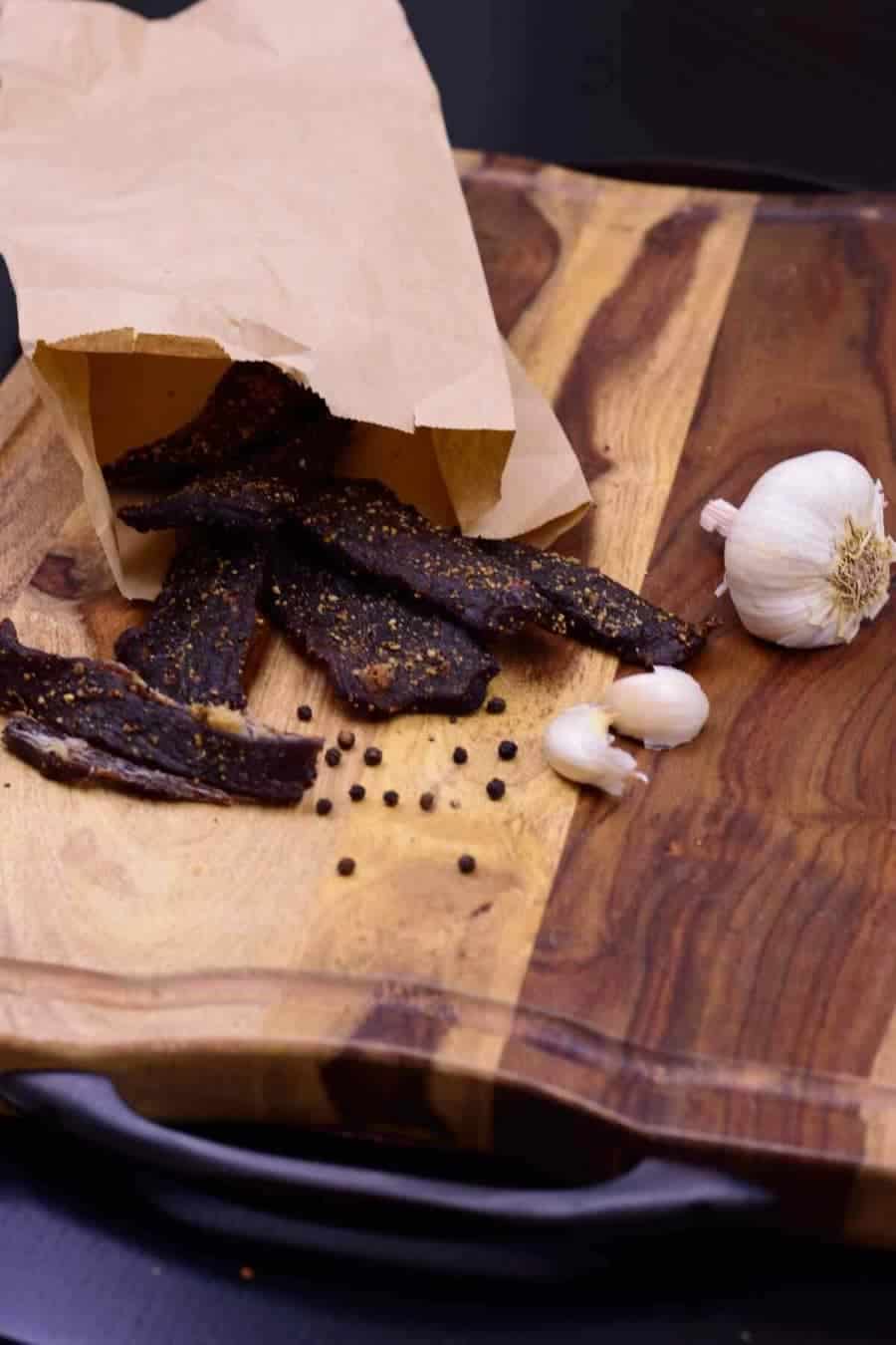 Garlic black pepper beef jerky in paper bag on cutting board with black peppercorns and garlic