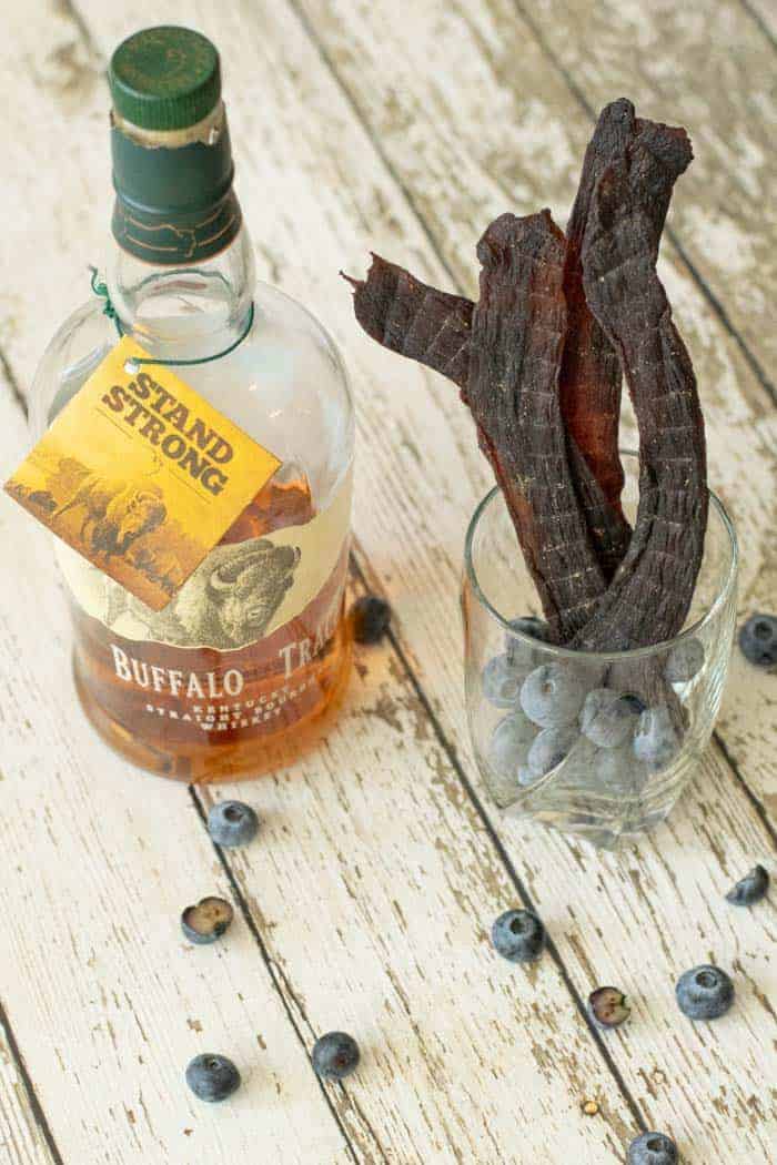 Blueberry deer jerky in a glass full of blueberries on wood table with a bottle of bourbon