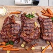 Three ribeye steaks cooked on cutting board with carrots and potatoes