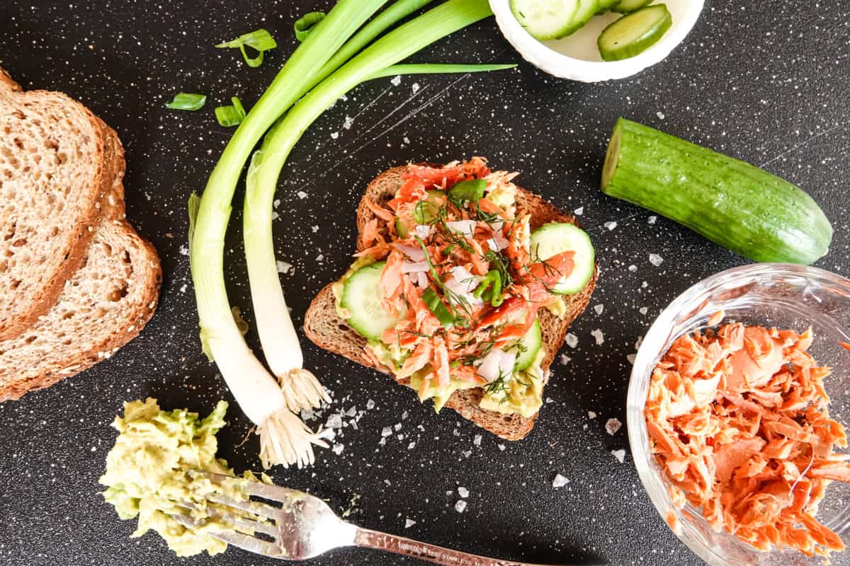 salmon avocado toast with bowl of salmon, cucumber, bread, and green onion on black cutting board