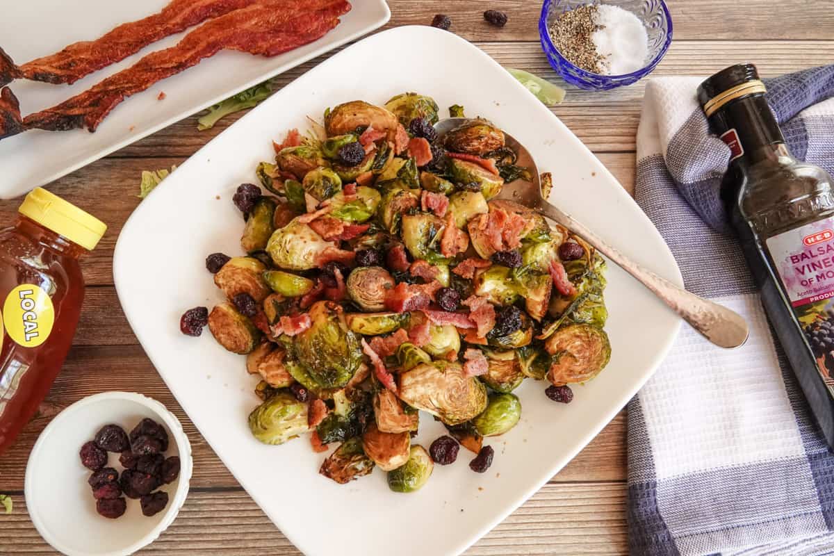Cooked brussel sprouts on white plate topped with bacon and cranberries.