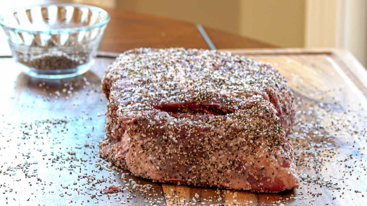Beef chuck roast covered in salt and pepper on cutting board with seasoning dish