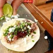 Sliced lime squeezed on pulled pork taco on white corn tortilla topped with chopped onion and jalapeño, cilantro