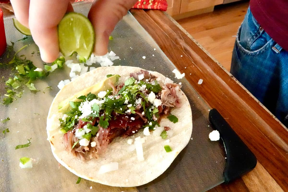 lime slice squeeze by hand, on top of smoked pull pork on a white corn tortilla garnished with crumbled white cheese, chopped up white onion and jalapeño, chopped cilantro and a slice of avocado on top of a glass board. 