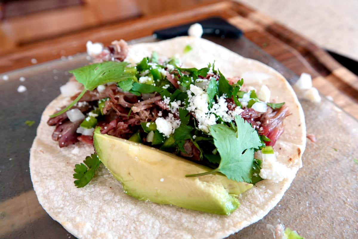 White corn tortilla topped with shredded smoked pork and chopped white onion and jalapeños, white crumbled cheese and cilantro leaves, and a slice of avocado  on glass cutting board with wood cutting board underneath.