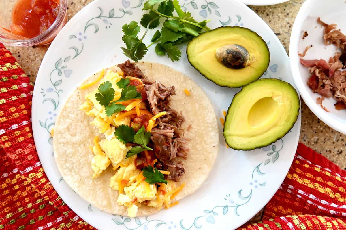 pulled pork, scrambled eggs, cilantro on taco with sliced avocado on white plate
