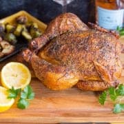 Smoked whole chicken on cutting board with lemons. Potatoes and brussel sprouts in background.