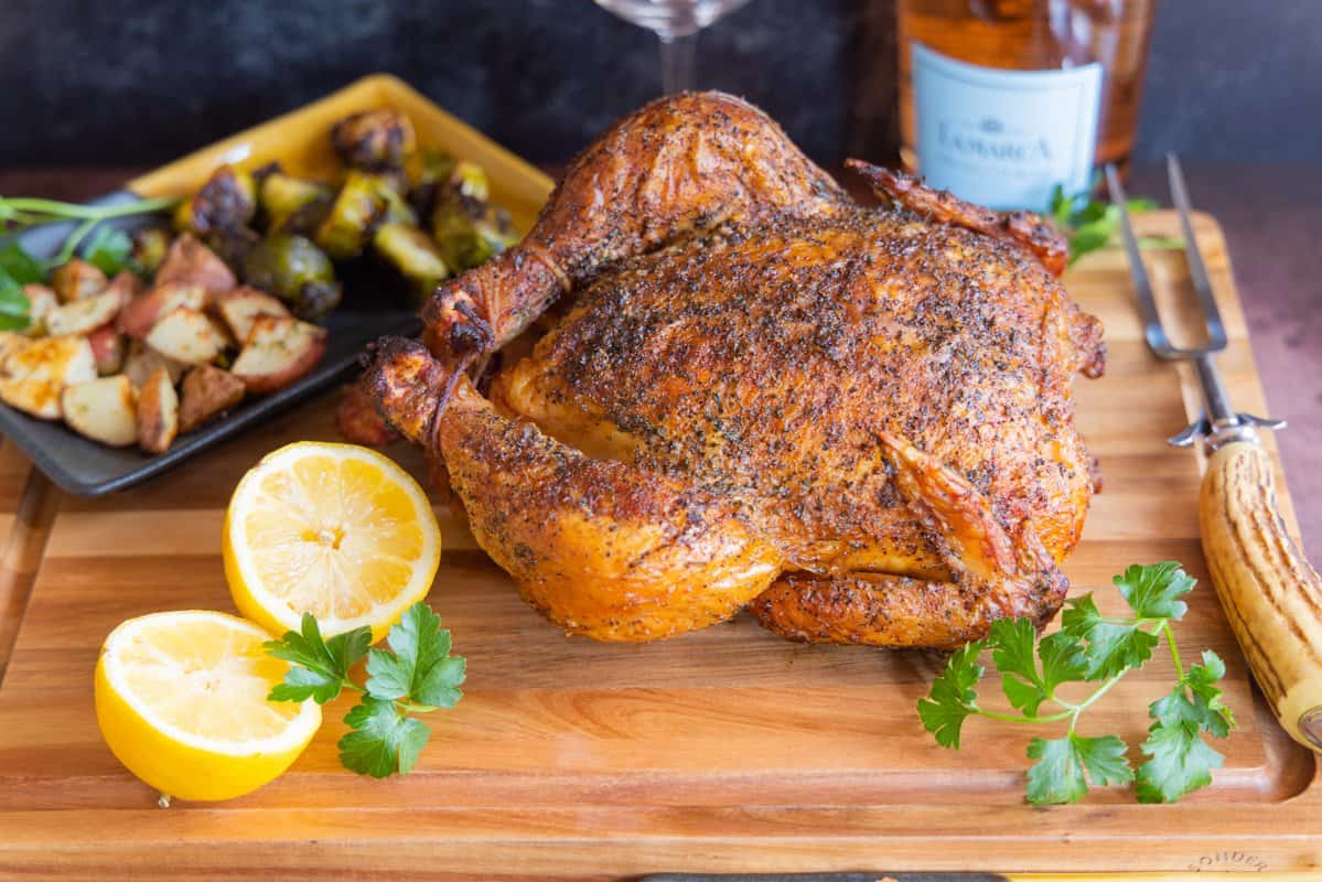 Smoked whole chicken on cutting board with lemons. Potatoes and brussel sprouts in background.