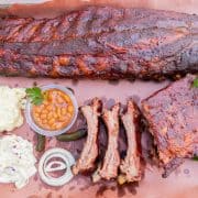 Rack of ribs and several individual ribs on butcher paper with coleslaw, potato salad, beans, and onions.
