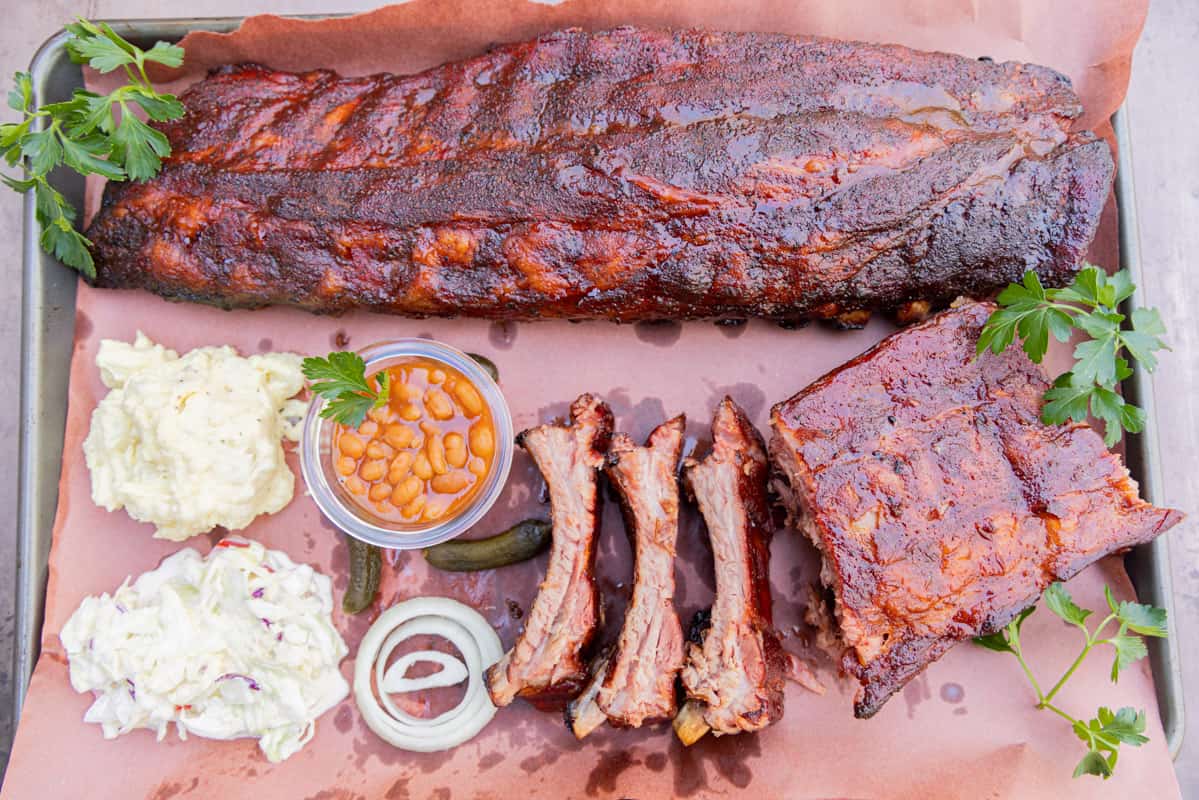 Rack of ribs and several individual ribs on butcher paper with coleslaw, potato salad, beans, and onions.
