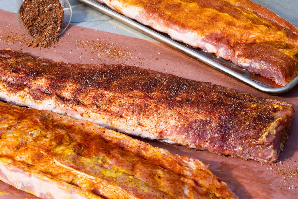 Racks of pork ribs seasoned with a dry rub sitting on butcher paper with spices in a glass jar.