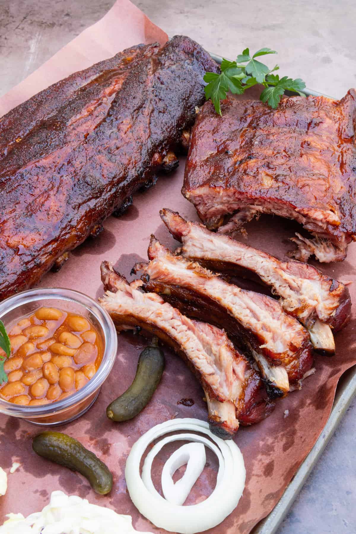 Rack of ribs on platter with onion, baked beans, pickles, and garnish.