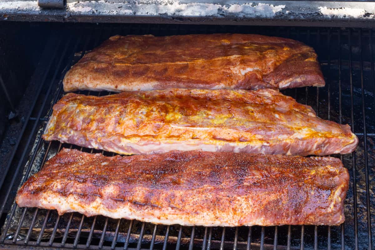 Three racks of pork ribs seasoned and beginning to smoke inside smoker on grill grates.