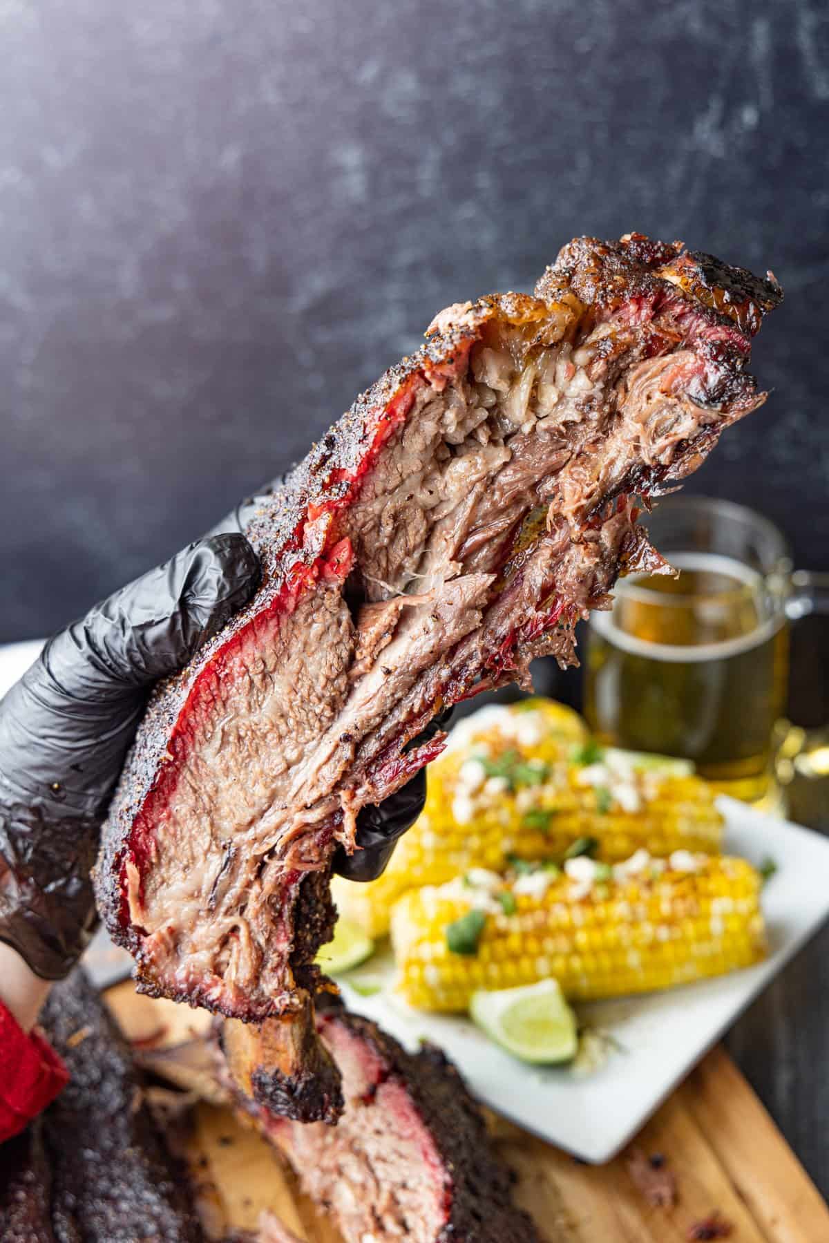 A large beef rib held in hand with corn and beer in background.