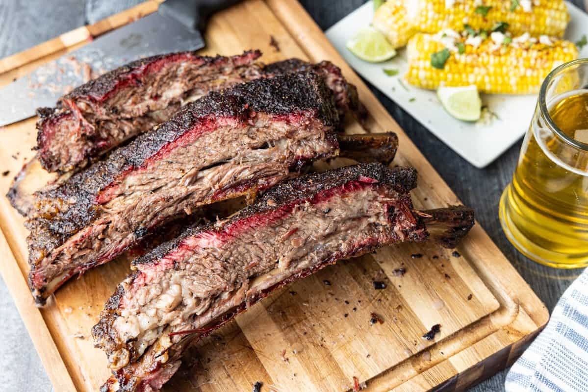 Beef ribs sliced on cutting board with corn on the cob and beer in a mug in the background.