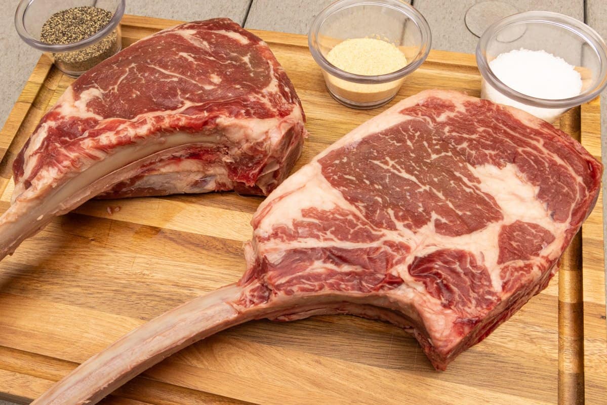 Two ribeye steaks on cutting board with salt, pepper, garlic in dishes.