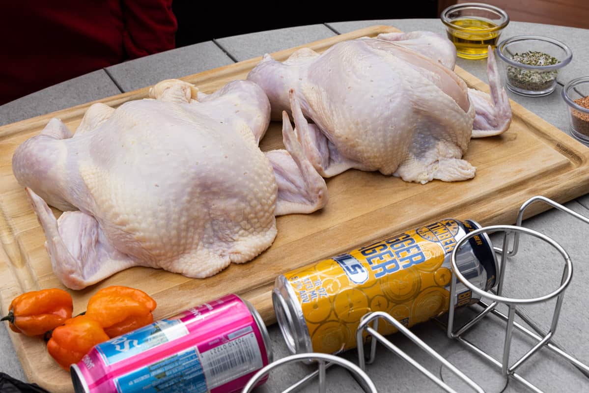 Two raw chickens on cutting board with beer cans and seasoning around the cutting board.