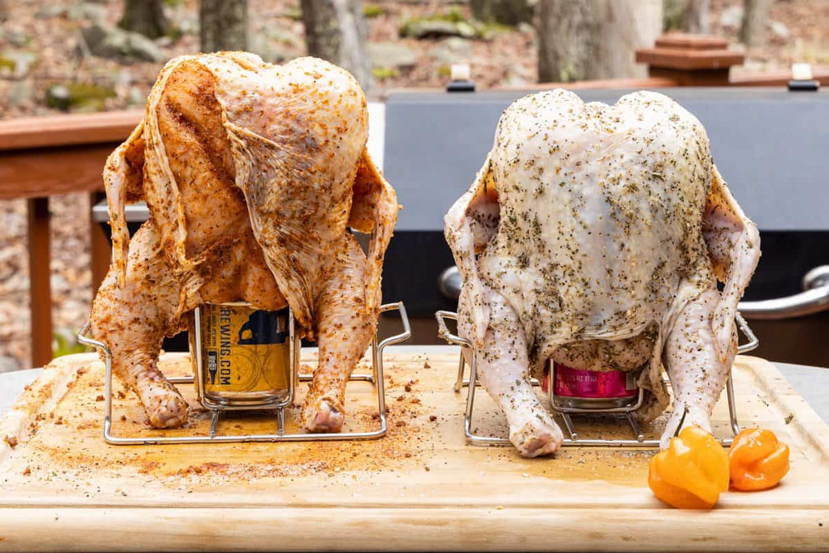 Two chickens seasoned and sitting atop beer cans on cutting board.