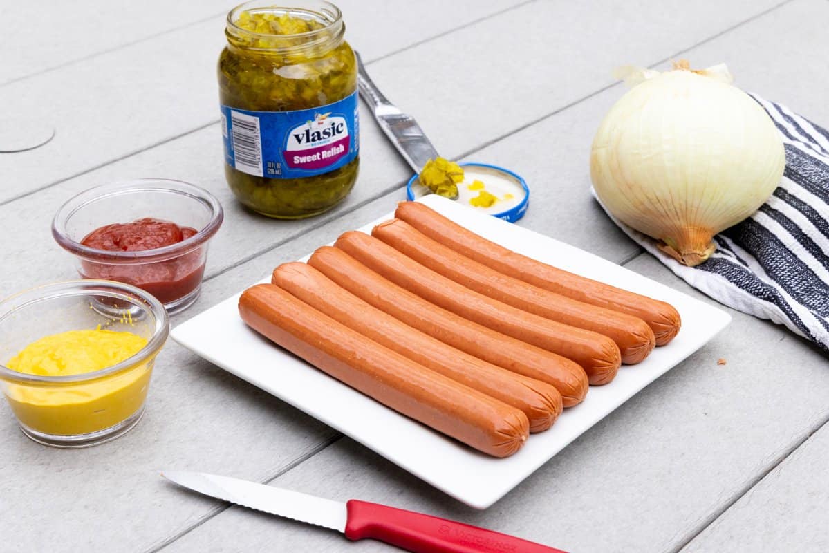 Hot dogs on a white plate with mustard, ketchup, relish, and onions around plate. 