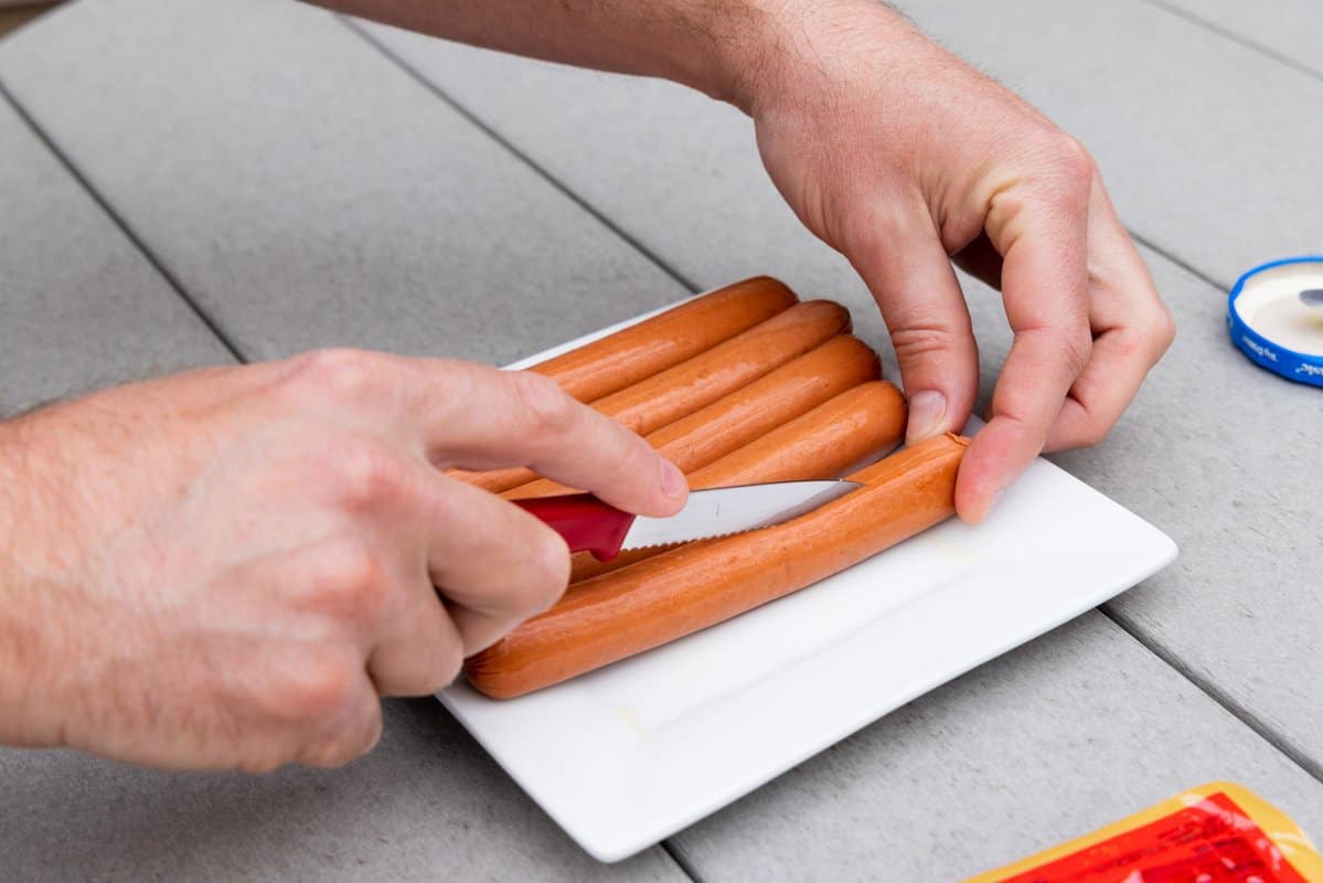 Slicing hot dogs down the middle with a sharp knife atop a white plate. 