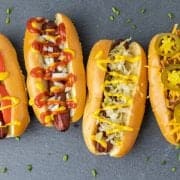 Four smoked hot dogs with different toppings on grey slate.