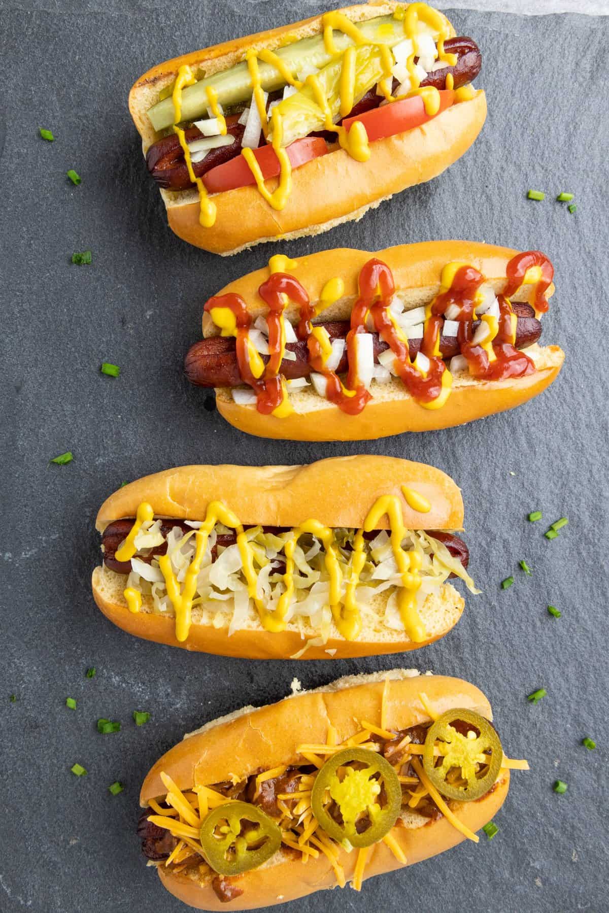 Four smoked hot dogs on grey slate with different toppings