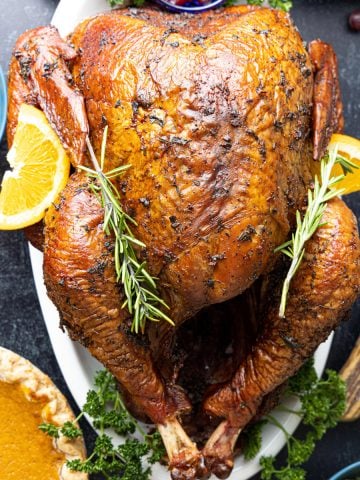 Turkey on white platter with orange slices as garnish with pumpkin pie and stuffing in background.