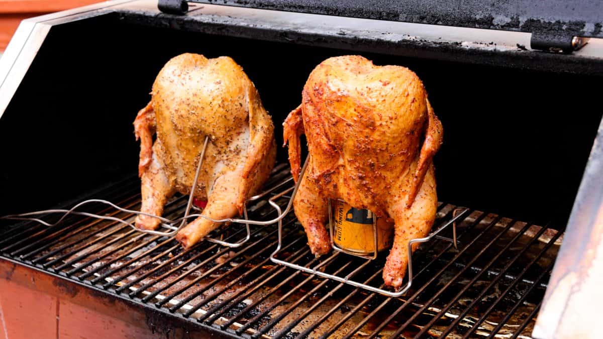 Two chickens roasting on grill standing up straight with beer can in cavity of chicken.