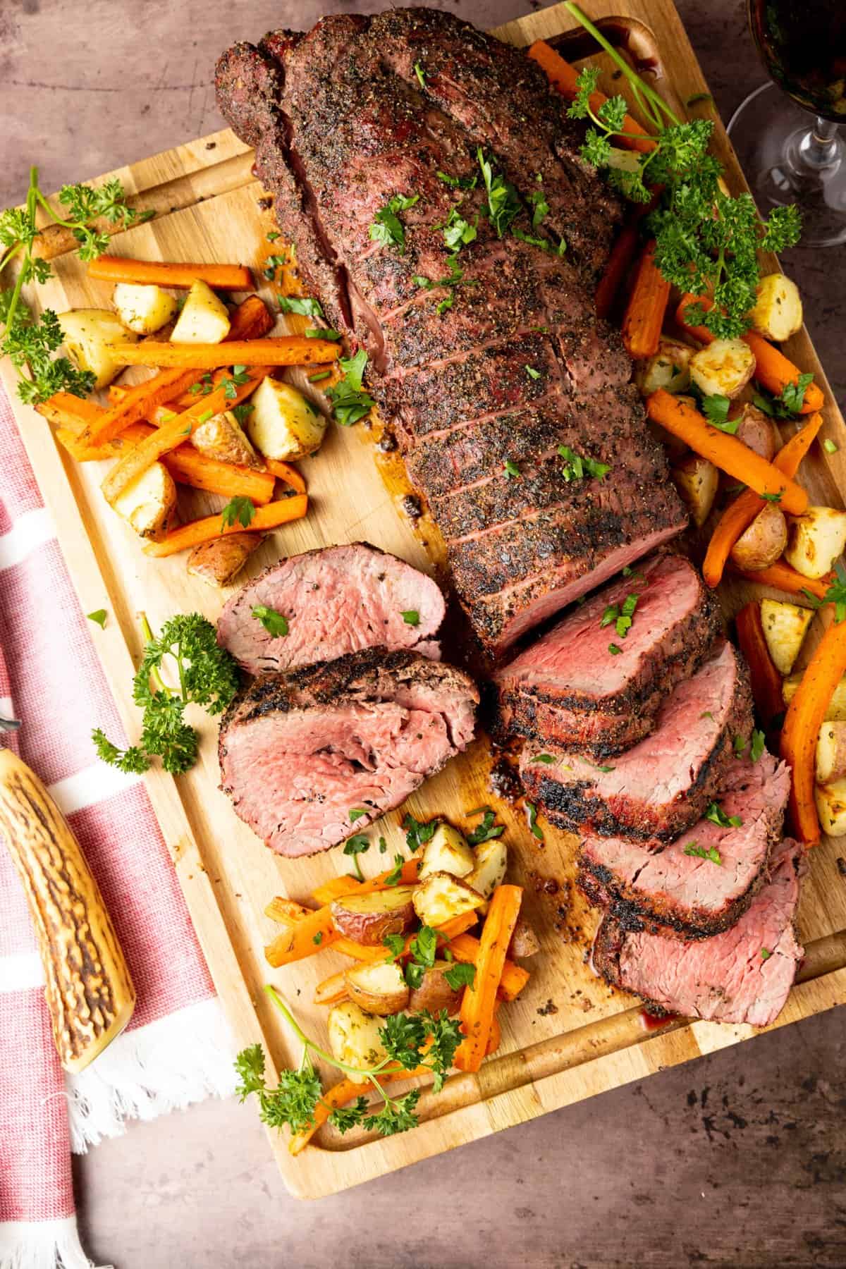 Smoked beef tenderloin on wood cutting board sliced to serve with potatoes and carrots surrounding the meat.