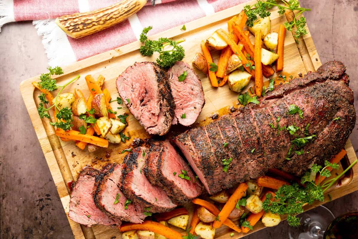 Beef tenderloin sliced on cutting board with potatoes and carrots surrounding meat.