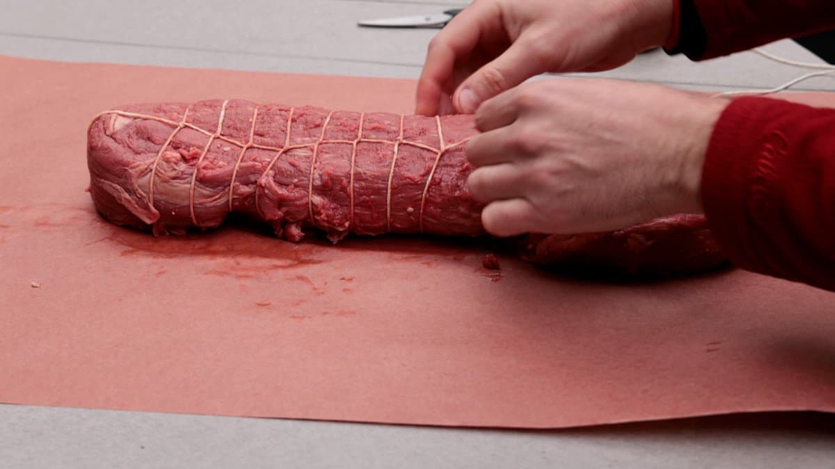 Meat roast on butcher paper being trussed with butcher twine.