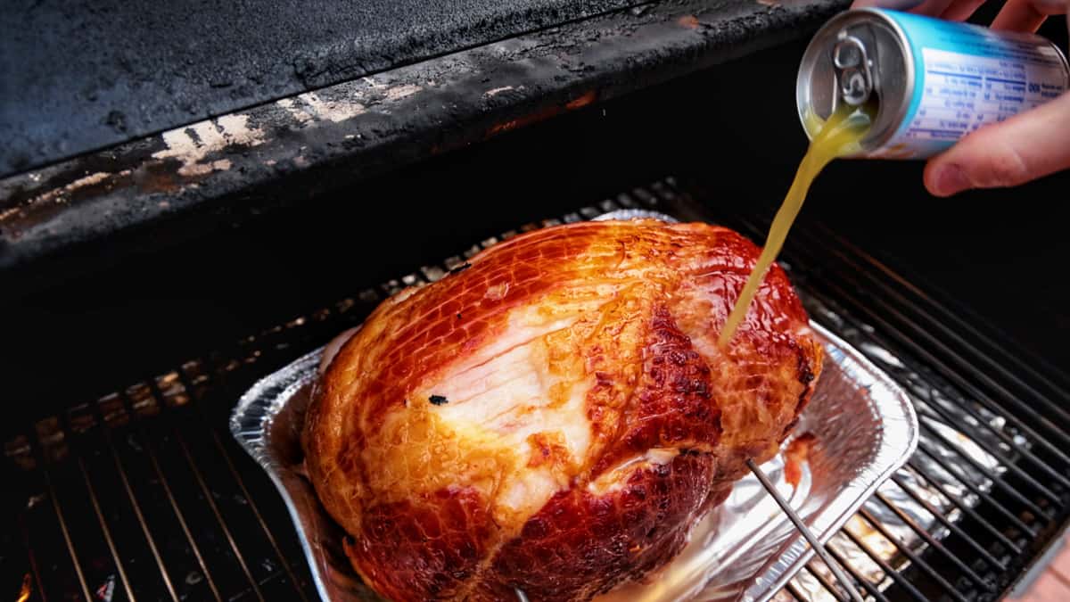 Pouring pineapple juice from a can over a ham that is on a smoker.