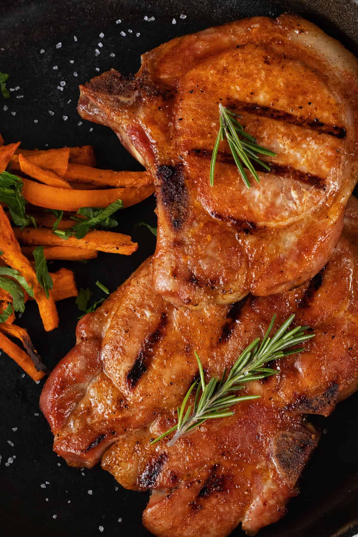 Two grilled pork chops on a black plate with carrots and rosemary garnish