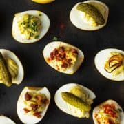 Deviled eggs on black platter with pickles, bacon, shallots, and paprika garnish.