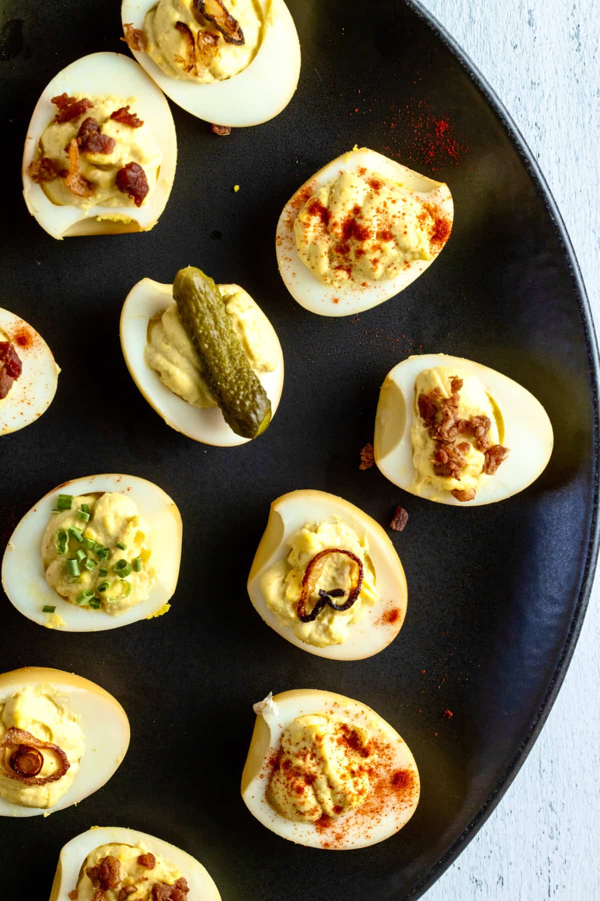 Deviled eggs on black platter with toppings such as pickles, bacon, paprika