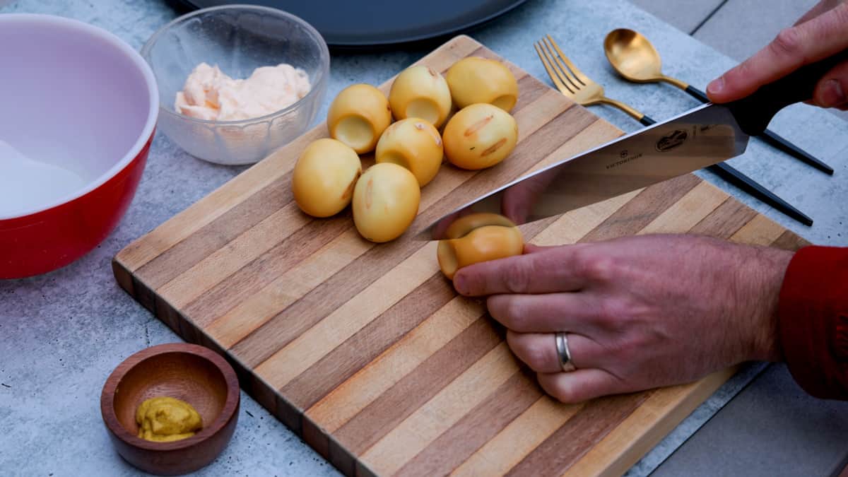 Slicing smoked eggs in half on cutting board with small dishes of mayonnaise and mustard nearby.