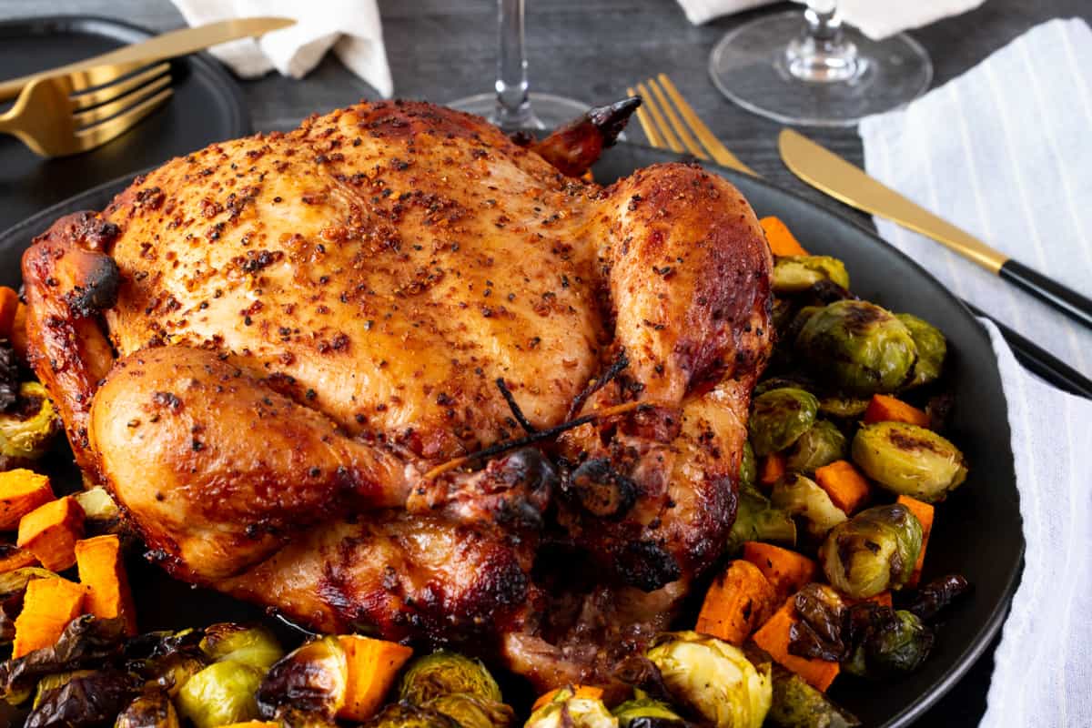 Cooked whole chicken on dark platter surrounded by vegetables and cutlery.