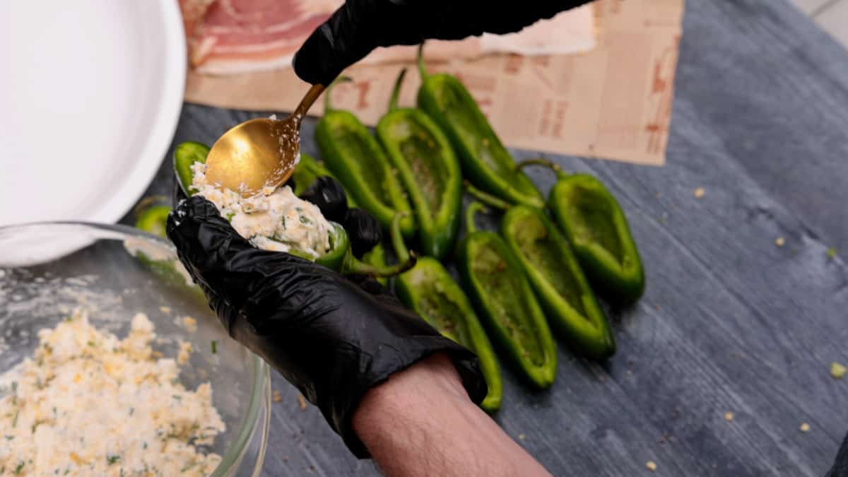 Filling jalapenos sliced in half with cream cheese with bacon in background on wood table.