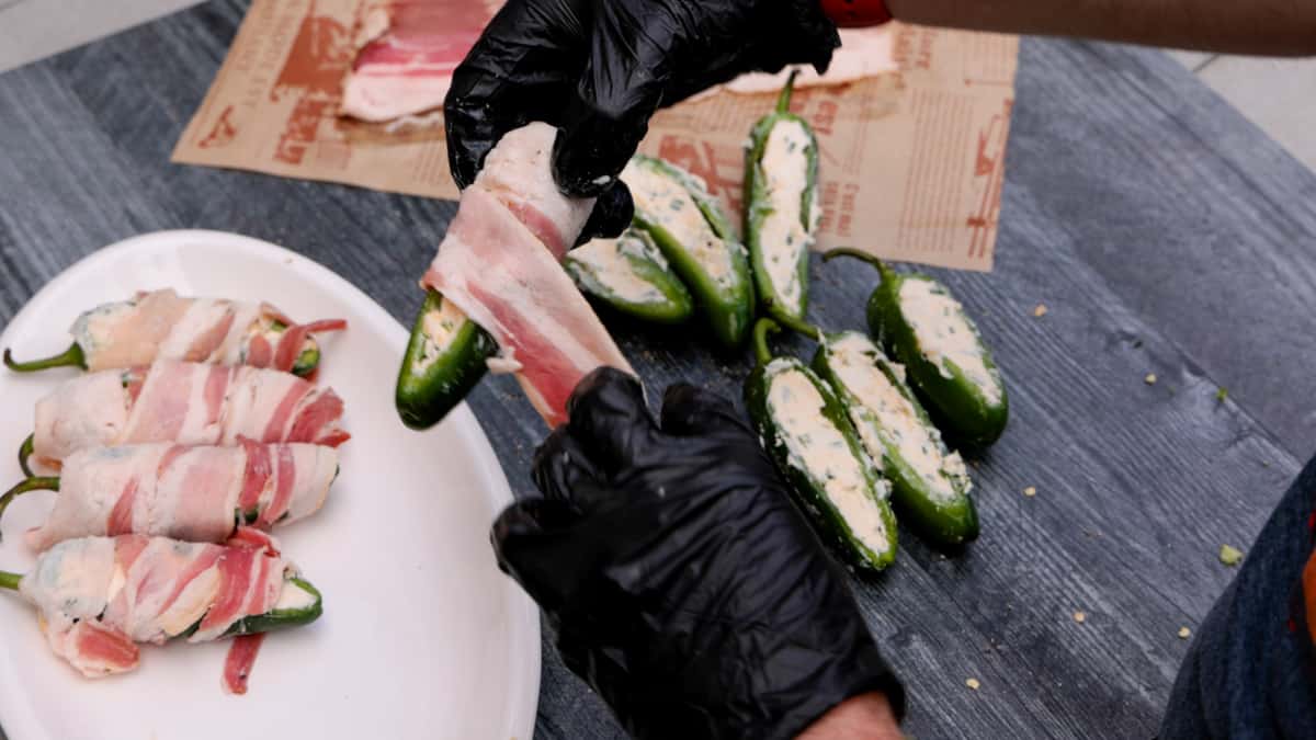 Wrapping a slice of bacon around a cream cheese stuffed jalapeno with a white plate in background with three wrapped jalapenos.
