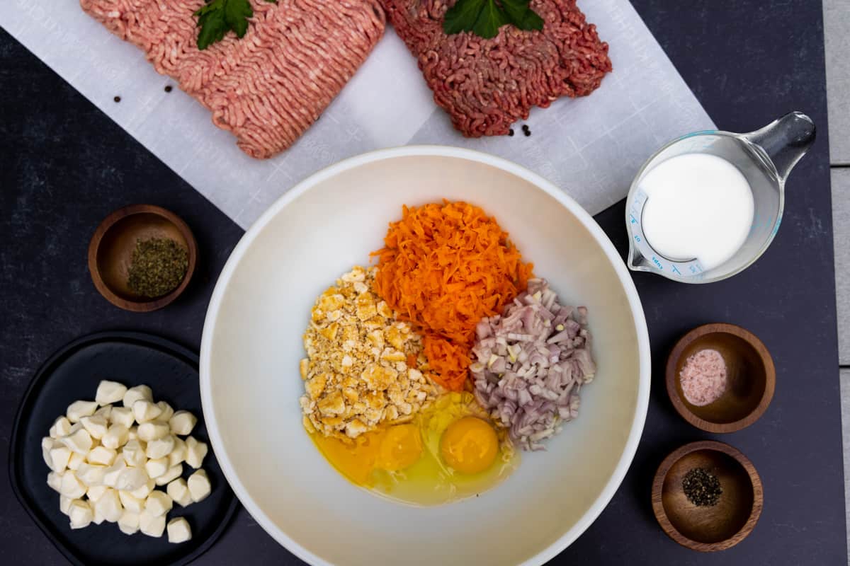 Meatloaf ingredients in a white bowl with ground pork and beef on white plates on the side.