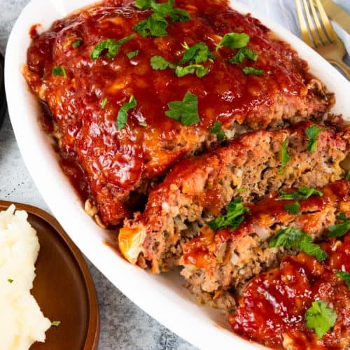 Meatloaf on white platter with sides of cabbage and mashed potatoes on wooden plates.
