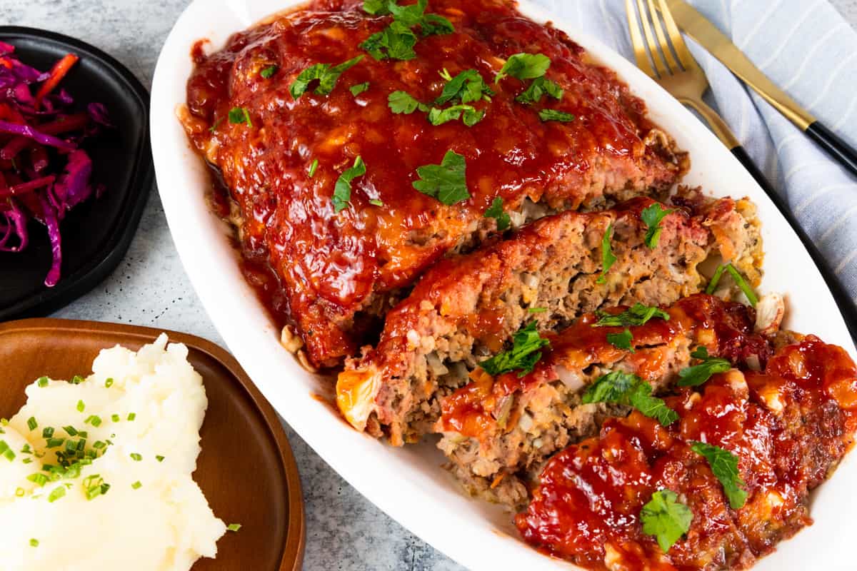 Meatloaf on white platter with sides of cabbage and mashed potatoes on wooden plates. 