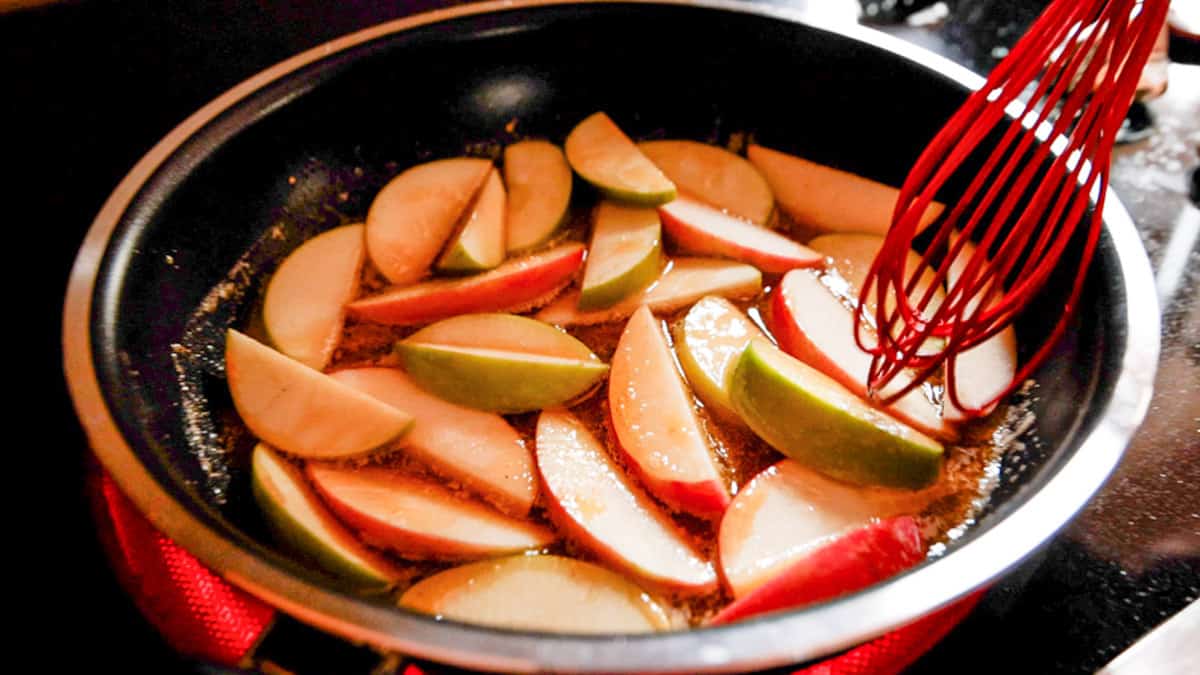 Sliced apples cooking in frying pan being stirred by a red whisk. 