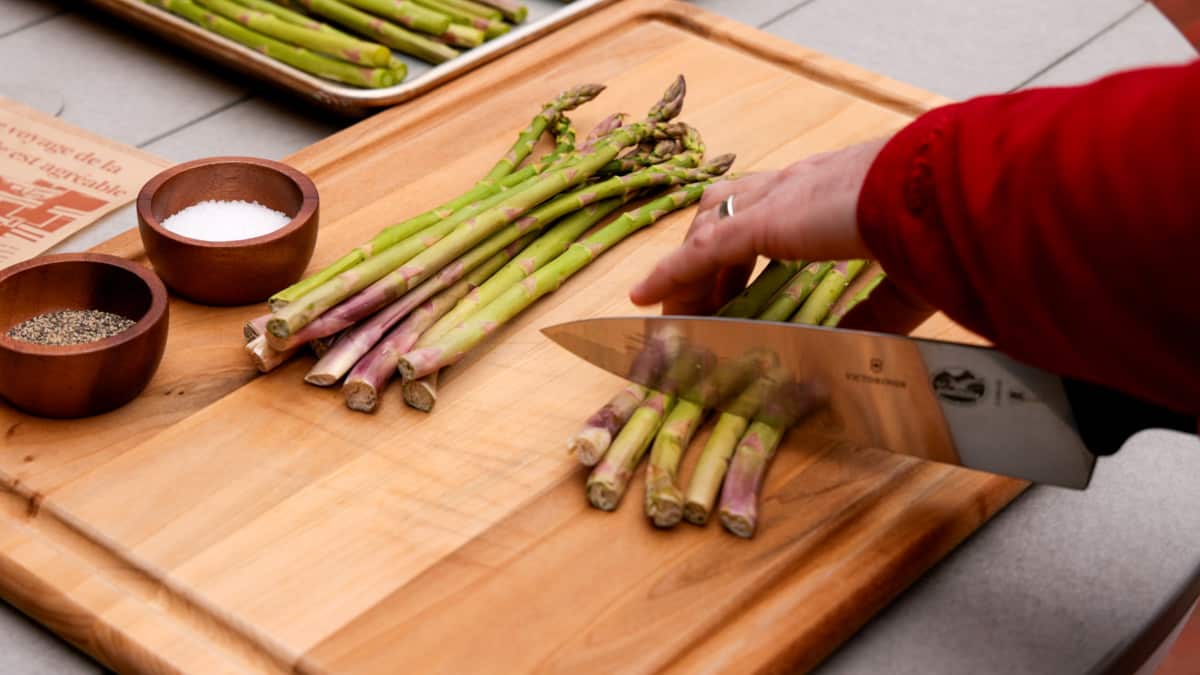 Cutting the ends off asparagus with a knife atop a wood cutting board.