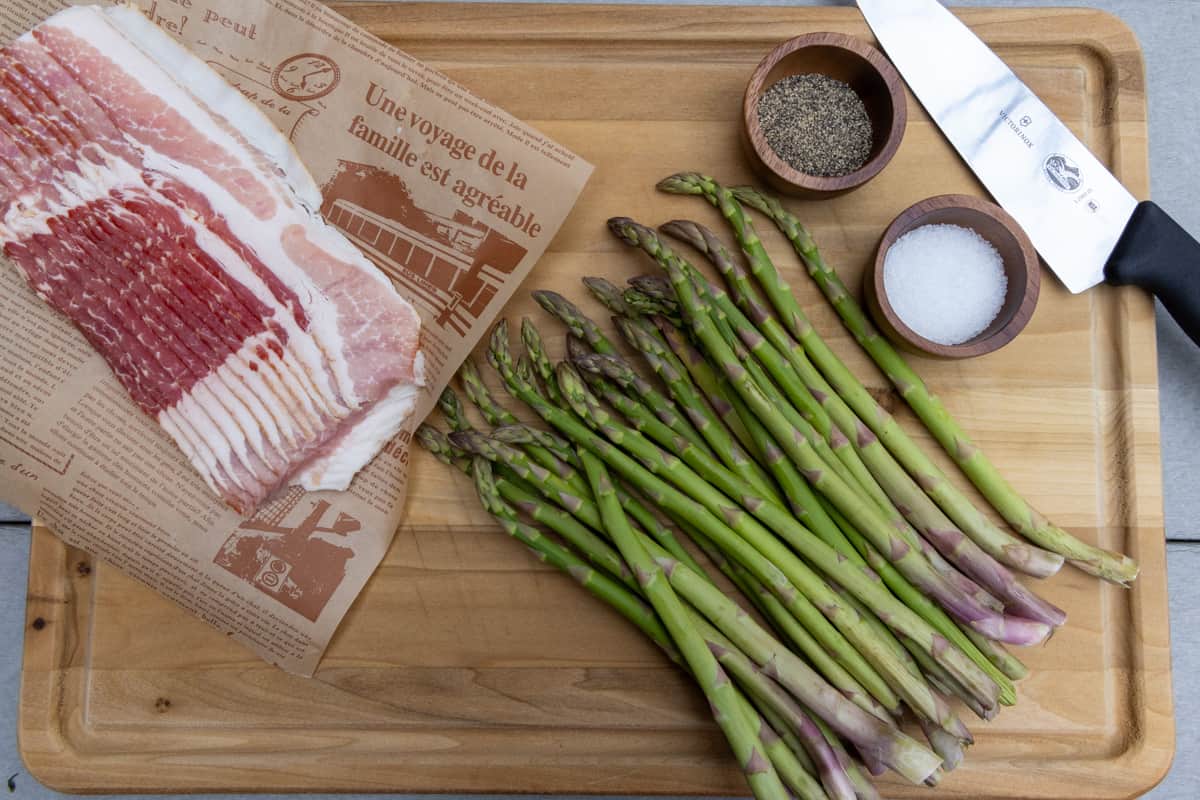 Asparagus, salt, black pepper, bacon, and a knife on a wood cutting board.
