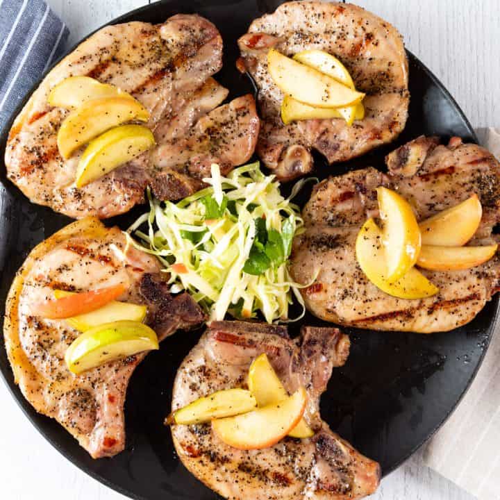 Black plate with 5 grilled pork chops topped with apples.