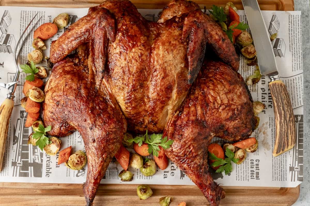 Spatchcock smoked turkey on cutting board atop butcher paper with vegetables and a carving knife and fork surrounding bird.
