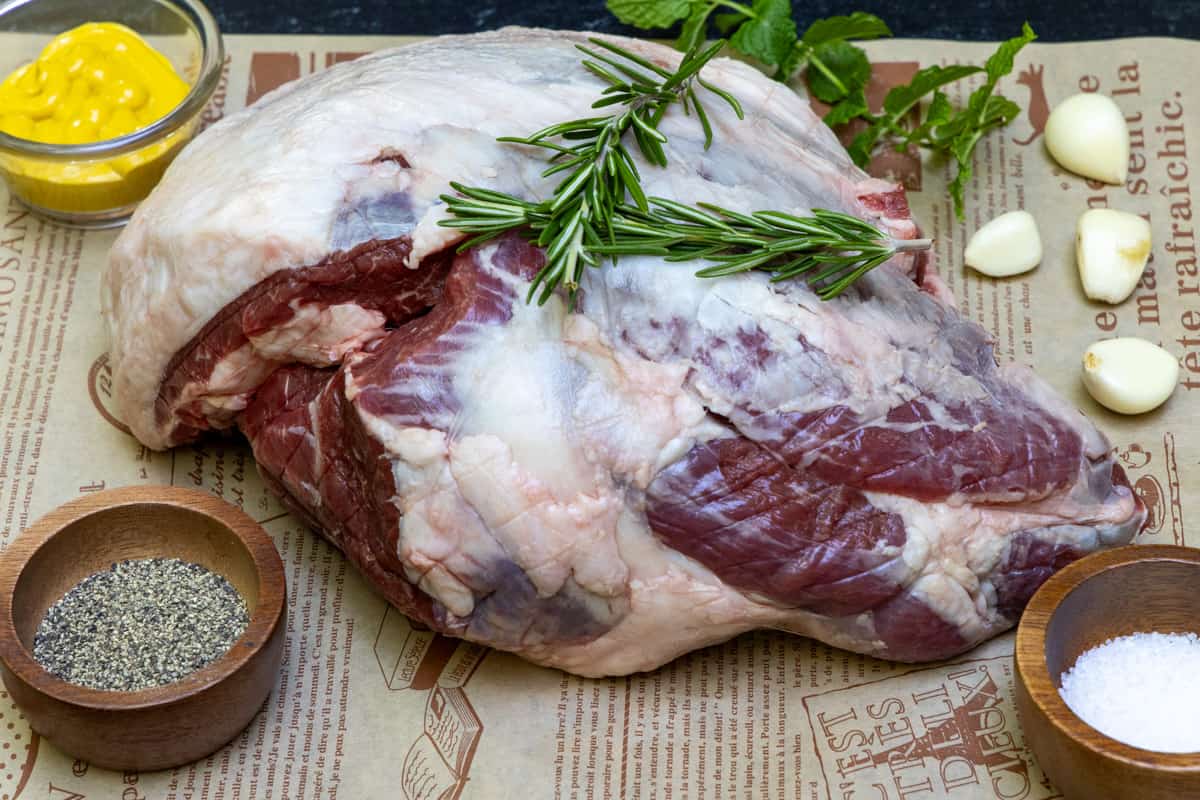 Leg of lamb on cutting board with pepper, mustard, salt, and garlic cloves surrounding meat.