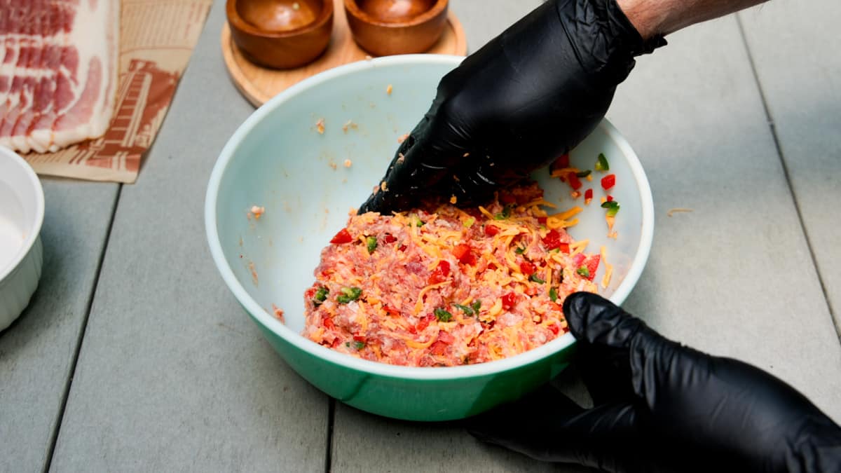 Mixing ground meat with cheese, peppers, and jalapeños in blue bowl by hand while wearing black gloves.