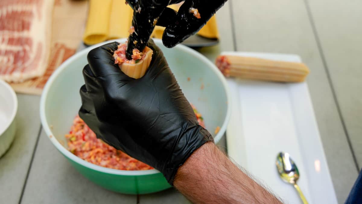 Stuffing meat mixture into manicotti shells by hand while wearing black gloves.